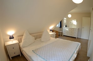 A bedroom with a sloped ceiling, a bed with white bedding, a nightstand with a lamp, a dresser, and a small table with a chair. - Vacation home with a fireplace and sauna, about 250 meters from the sandy beach, Wi-Fi, washing machine (Glowe)