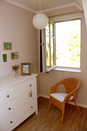 Ein Zimmer mit weißem Schminktisch, Korbstuhl und einem Fenster mit Jalousien.