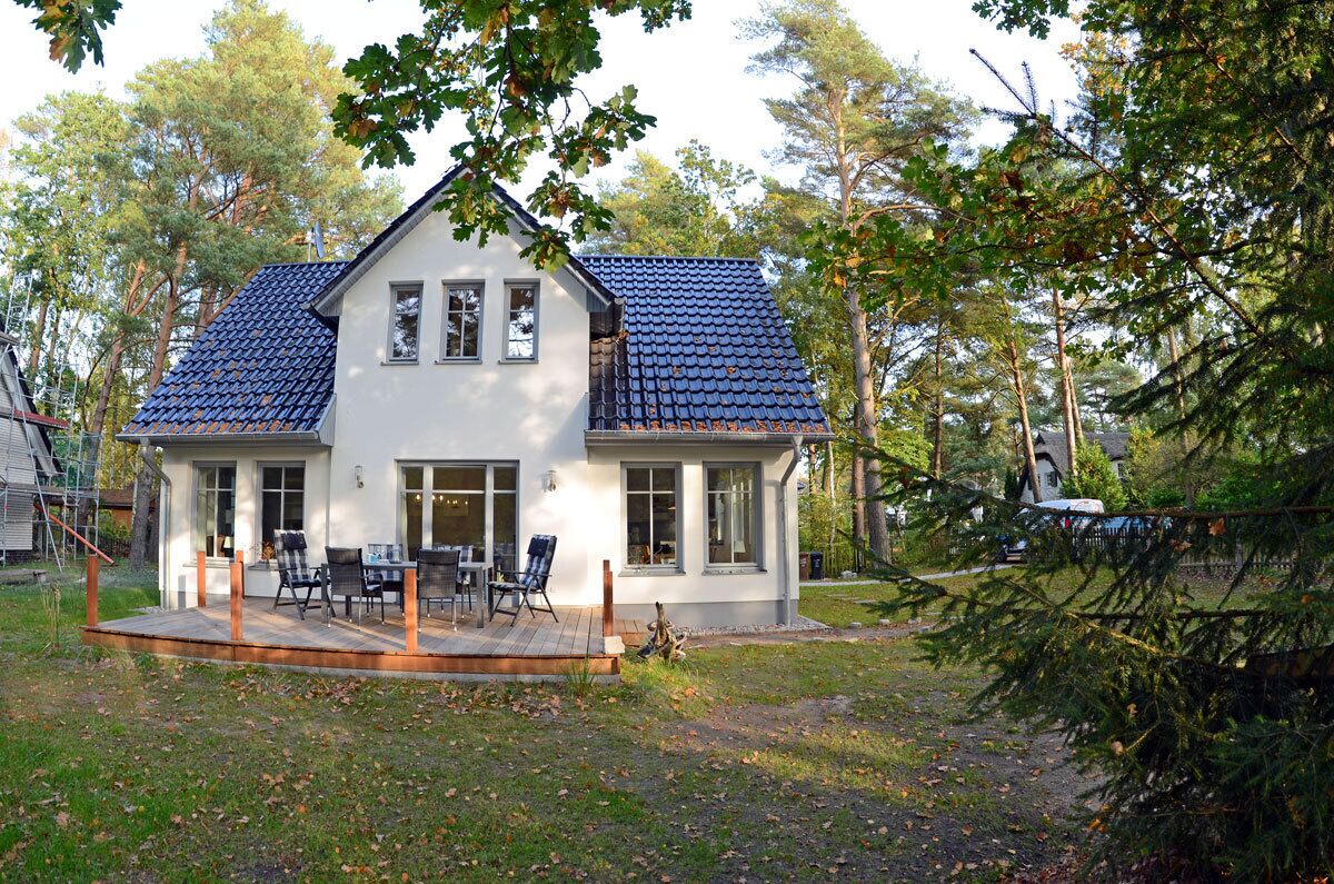 A two-story house with a tiled roof, a covered patio with outdoor furniture, and a surrounding wooded area.