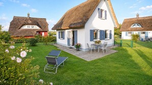Une maison avec un toit de chaume, une terrasse avec une table et des chaises, et une pelouse bien entretenue.