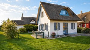 Une maison avec un toit de chaume, un petit jardin avec une pelouse et une clôture, et une table avec des chaises à l’extérieur.