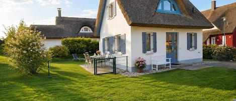 A house with a thatched roof, a small garden with a lawn and a fence, and a table with chairs outside.