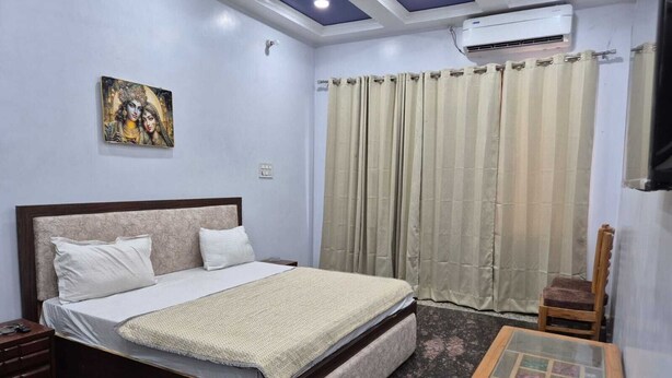 A bedroom with a bed, a wooden nightstand, a glass-top table, a chair, a ceiling fan, and a painting on the wall. - Prince Hotel (Meerut)