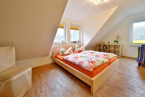 A bedroom with a sloped ceiling, a bed with a floral bedspread, a bedside table, a chair, and a window with a view. - Vacation home with a fireplace, about 350 meters from the sandy beach, pets welcome (Glowe)