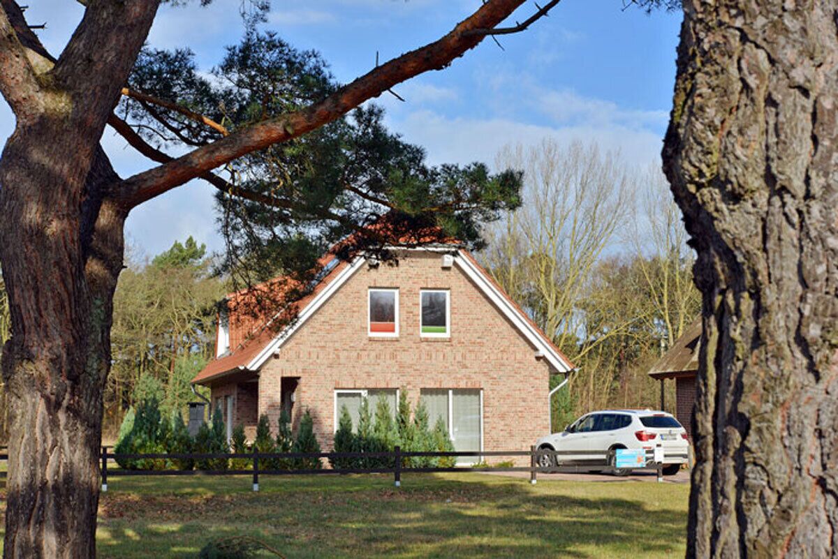 A brick house with a gabled roof, surrounded by trees and a parked car.