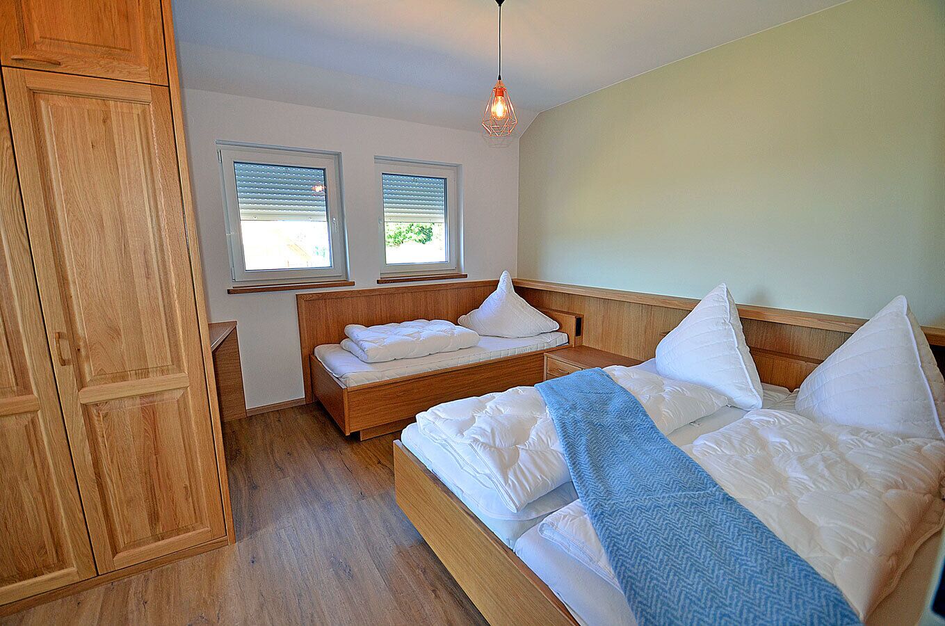 Ein Schlafzimmer mit einem hölzernen Bett, zwei Fenstern mit Jalousien, einem Kleiderschrank und einer Pendelleuchte.