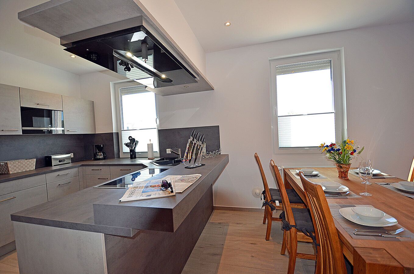 A modern kitchen with a central island, dining area with wooden chairs, and large windows providing natural light.