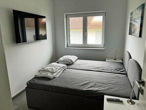 Een slaapkamer met een bed, nachtkastje, wandtelevisie en raam.