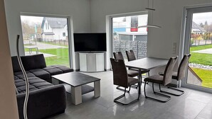 Una sala de estar moderna con un sofá, una mesa de centro y una mesa de comedor con sillas. Hay un televisor de pantalla plana montado en la pared.