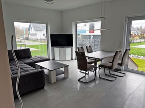 A modern living room with a sofa, a coffee table, and a dining table with chairs. There is a flat-screen TV mounted on the wall. - Modern vacation rental with a fireplace, about 1,300 meters from the sandy beach, 2 bedrooms, sleeps up to 5 people (Glowe)