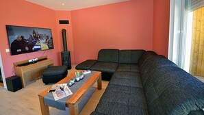 A living room with a large sectional sofa, a television mounted on the wall, and a small wooden table with magazines and a plant. - 2-story vacation home, quiet location, about 1.3 kilometers from the sandy beach (Baltic Sea) (Glowe)