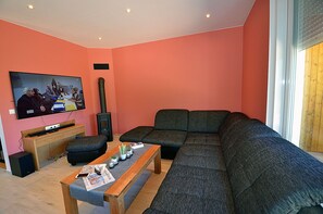 Ein Wohnzimmer mit einem großen Ecksofa, einem an der Wand montierten Fernseher und einem kleinen Holztisch mit Zeitschriften und einer Pflanze.