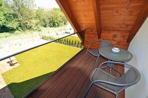 Un balcone con pavimento in legno, due sedie in vimini, un piccolo tavolino rotondo e vista su un prato verde e alberi.