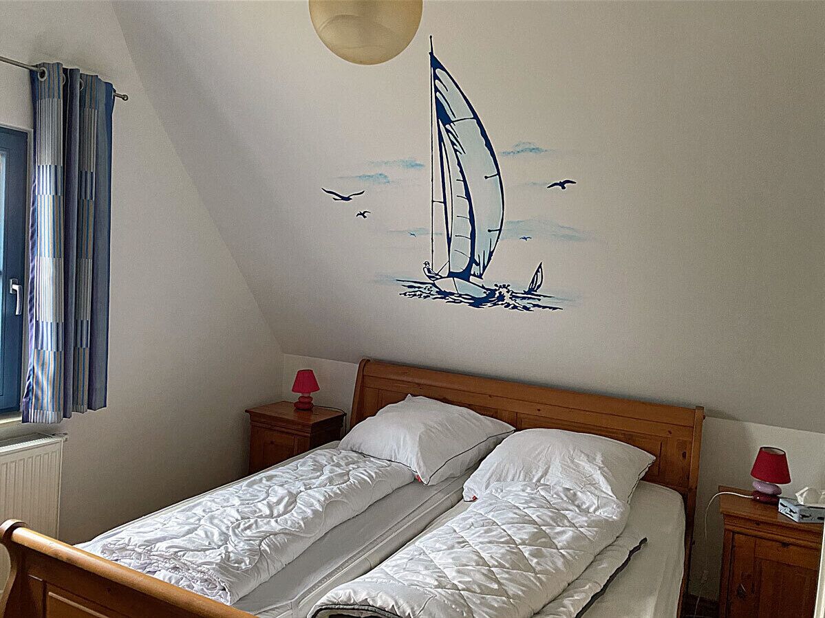 A bedroom with a single bed, two bedside tables, a window with curtains, and a wall mural of a sailboat.