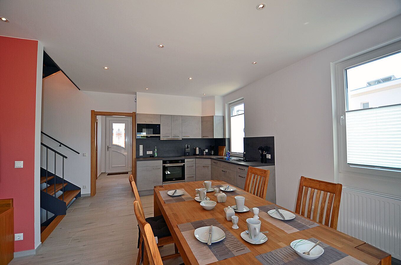 A modern kitchen with grey cabinets, a dining area with a wooden table set for four, and a staircase leading to an upper floor.