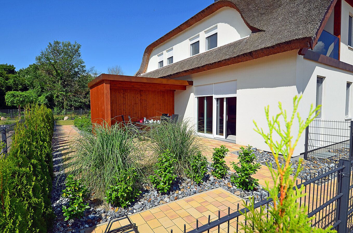 A modern house with a thatched roof, a covered outdoor seating area, and a garden with various plants and a paved walkway.