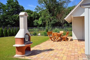 Un patio exterior con una parrilla de ladrillo, una mesa y sillas de madera, y un edificio blanco al fondo.