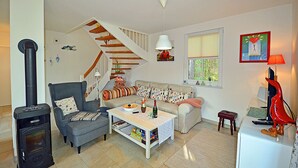 Een gezellige woonkamer met een bank, fauteuil en een tafel met een fles en glazen. Een trap met houten treden leidt naar een andere kamer.