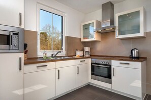 Een moderne keuken met witte kastjes, een oven van roestvrij staal, een magnetron en een raam dat voor natuurlijk licht zorgt.