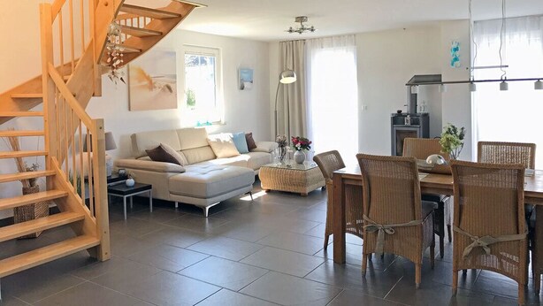 Ein modernes Wohnzimmer mit einer Holztreppe, einem Essbereich mit Korbstühlen und einer Küche mit einer hängenden Leuchte.