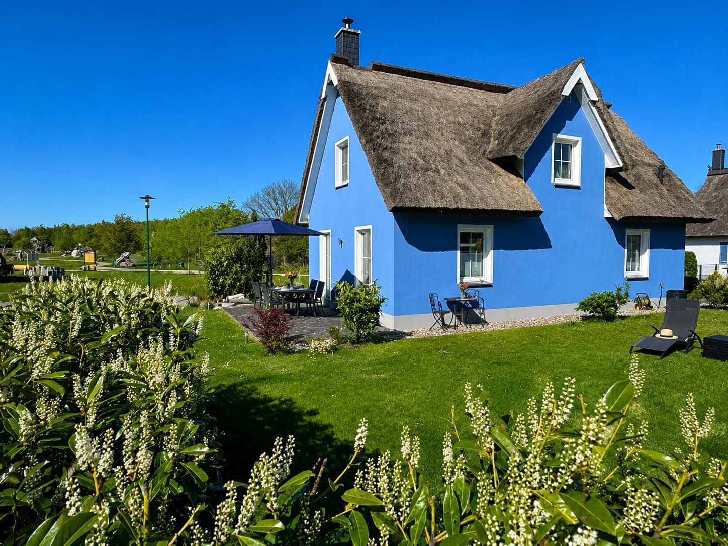 Une maison bleue avec un toit de chaume, une terrasse avec des meubles d’extérieur et une pelouse bien entretenue.