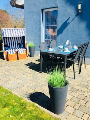 Een terras buiten met een eettafel, stoelen, een gestreepte zithoek en potplanten.