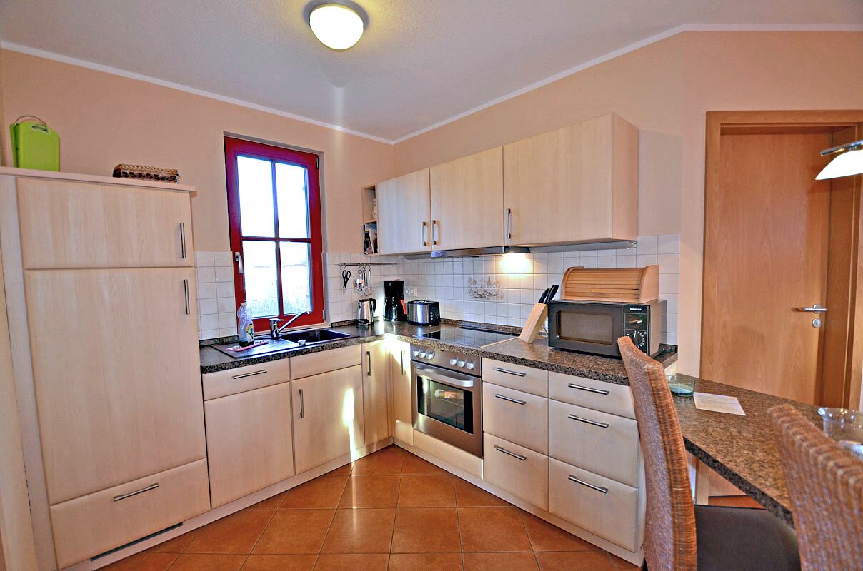 A kitchen with light wooden cabinets, a stainless steel oven, a microwave, and a window with a red frame.