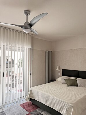 Een hotelkamer met een bed, een plafondventilator, een radiator en uitzicht door een raam met jaloezieën.