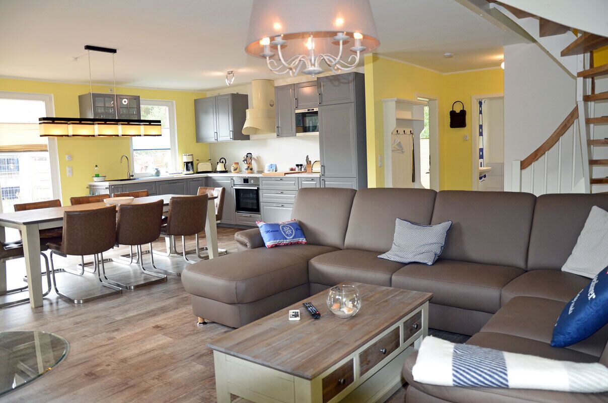 Een moderne woonkamer met een modulaire bank, een eethoek met tafel en stoelen, een keuken met grijze kasten, en een trap.