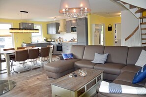 A modern living room with a sectional sofa, a dining area with a table and chairs, a kitchen with grey cabinets, and a staircase. - Vacation home with a fireplace and sauna, about 300 meters from the sandy beach; pets welcome (Glowe)