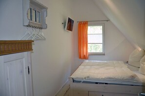 Une chambre avec un lit simple, une fenêtre avec des rideaux, une porte blanche et une étagère murale ornée d’un tableau.