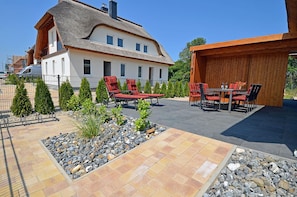 A house with a thatched roof, a patio with outdoor furniture, and a garden with small trees and a rock bed.
