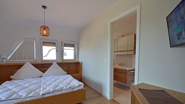 A bedroom with a wooden bed, a nightstand, a hanging light fixture, a window with blinds, and a view into the bathroom.