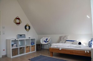 Une chambre avec un plafond en pente, un lit avec une literie blanche, une chaise ornée d’un coussin à motif d’ancre et une étagère blanche avec des paniers.