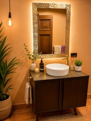Un baño con un lavamanos blanco redondo, un mueble de madera oscura y un espejo con un marco de mosaico. En la encimera, hay una planta en maceta, una vela y una botella de jabón de manos.