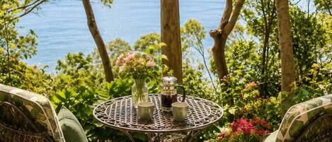 An outdoor patio with a round metal table, two wicker chairs with cushions, and a view of the sea.