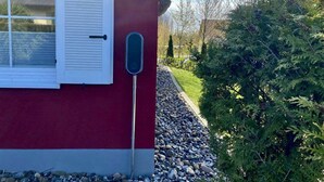 Un bâtiment rouge avec une fenêtre et des volets blancs, une boîte aux lettres en métal et un chemin de gravier menant à un buisson vert.