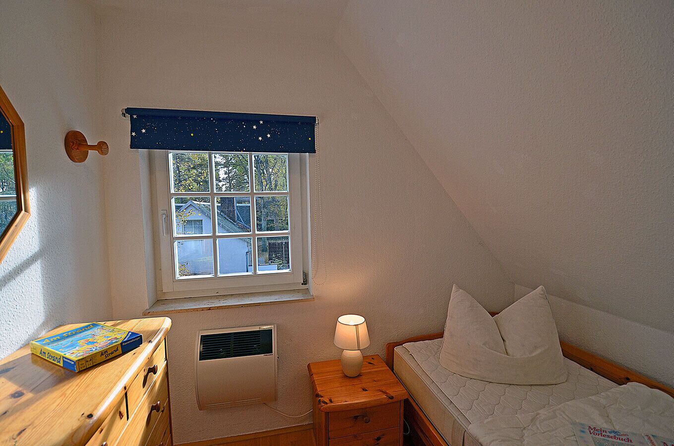 Une chambre avec une fenêtre, une table de chevet avec une lampe, un lit avec des oreillers et une commode en bois sur laquelle repose un livre.