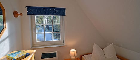 A bedroom with a window, a bedside table with a lamp, a bed with pillows, and a wooden dresser with a book.