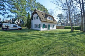 A white house with a thatched roof and green shutters, surrounded by a well-maintained lawn and trees. - Vacation home with a sauna, about 300 meters from the sandy beach, Wi-Fi, and a washing machine (Glowe)