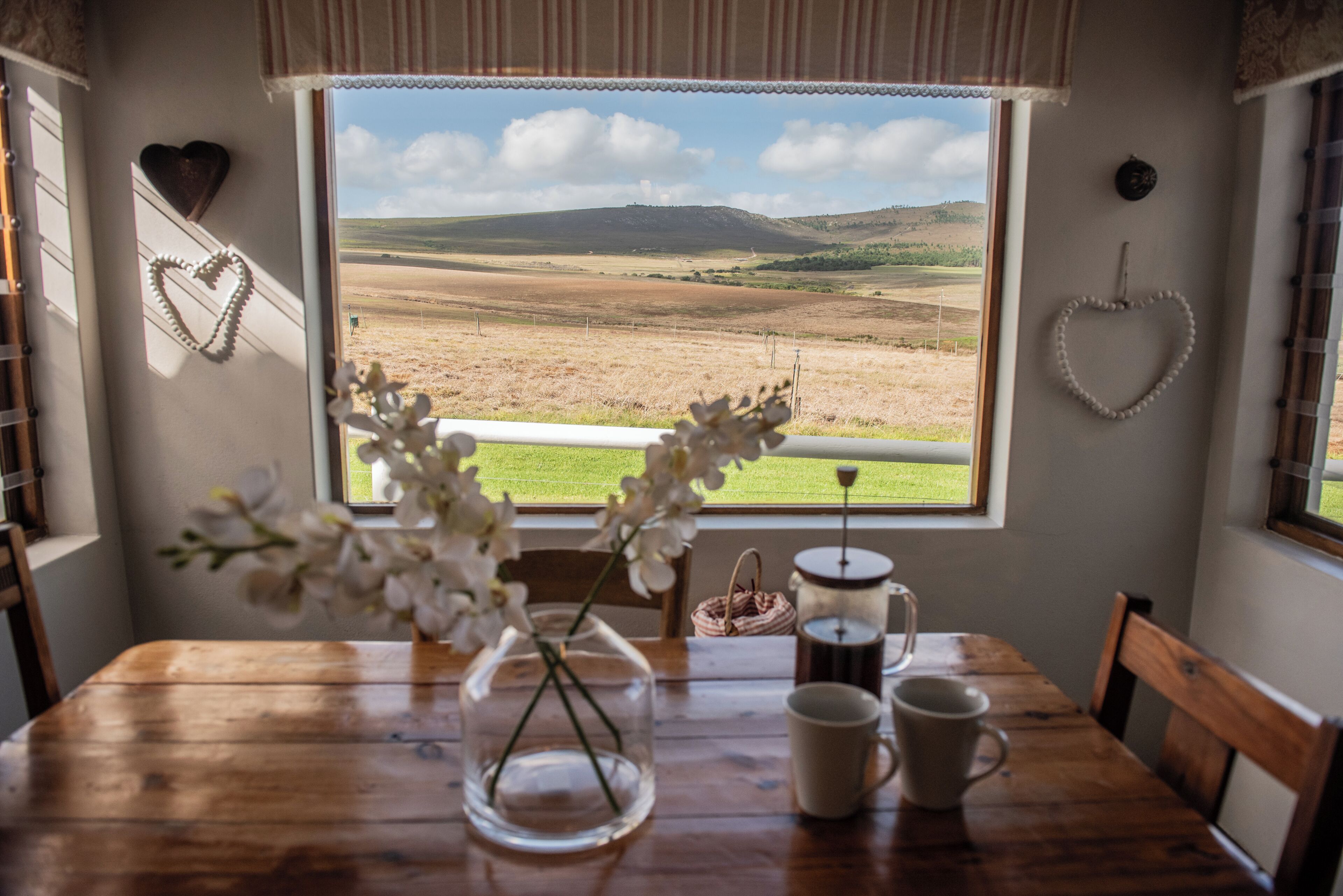 A room with a view of a landscape through a window, a wooden table with a vase of flowers, two cups, and a kettle.