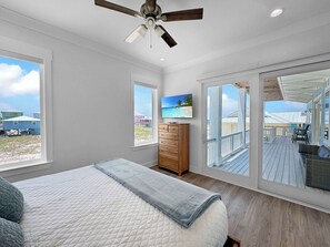 A bedroom with a bed, a dresser, a television, and a large window with a view of the beach and other buildings. - The Fort - Gulf Coast Getaway (Gulf Shores)