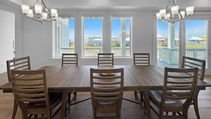 Ein Esszimmer mit großem Holztisch und sechs Stühlen, großen Fenstern mit Blick auf Gebäude und Pendelleuchten.