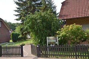 Une entrée fermée par un portail mène à une propriété comprenant un bâtiment en briques, un panneau indicateur et un grand arbre.