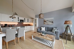 Eine moderne Küche mit weißen Schränken, ein Essbereich mit zwei Stühlen, einem Couchtisch und einem grauen Sofa in einem Wohnzimmer mit einem Bild an der Wand.