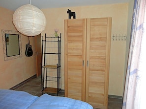 Ein Schlafzimmer mit einem Bett, einem Kleiderschrank, einer Hängelampe, einer Gitarre und einem kleinen Regal mit Blumen.