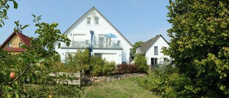 A two-story house with a balcony, surrounded by greenery and a stone wall.