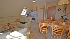 Een compacte woonruimte met een bank, een eettafel met stoelen, een kitchenette en een raam dat voor daglicht zorgt.