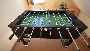 Een tafelvoetbaltafel met een voetbalveld-indeling, een raam met zonwering en een kleine tafel met een plant.