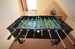 Ein Kicker mit Fußballfeld-Optik, ein Fenster mit Jalousien und ein kleiner Tisch mit einer Pflanze.
